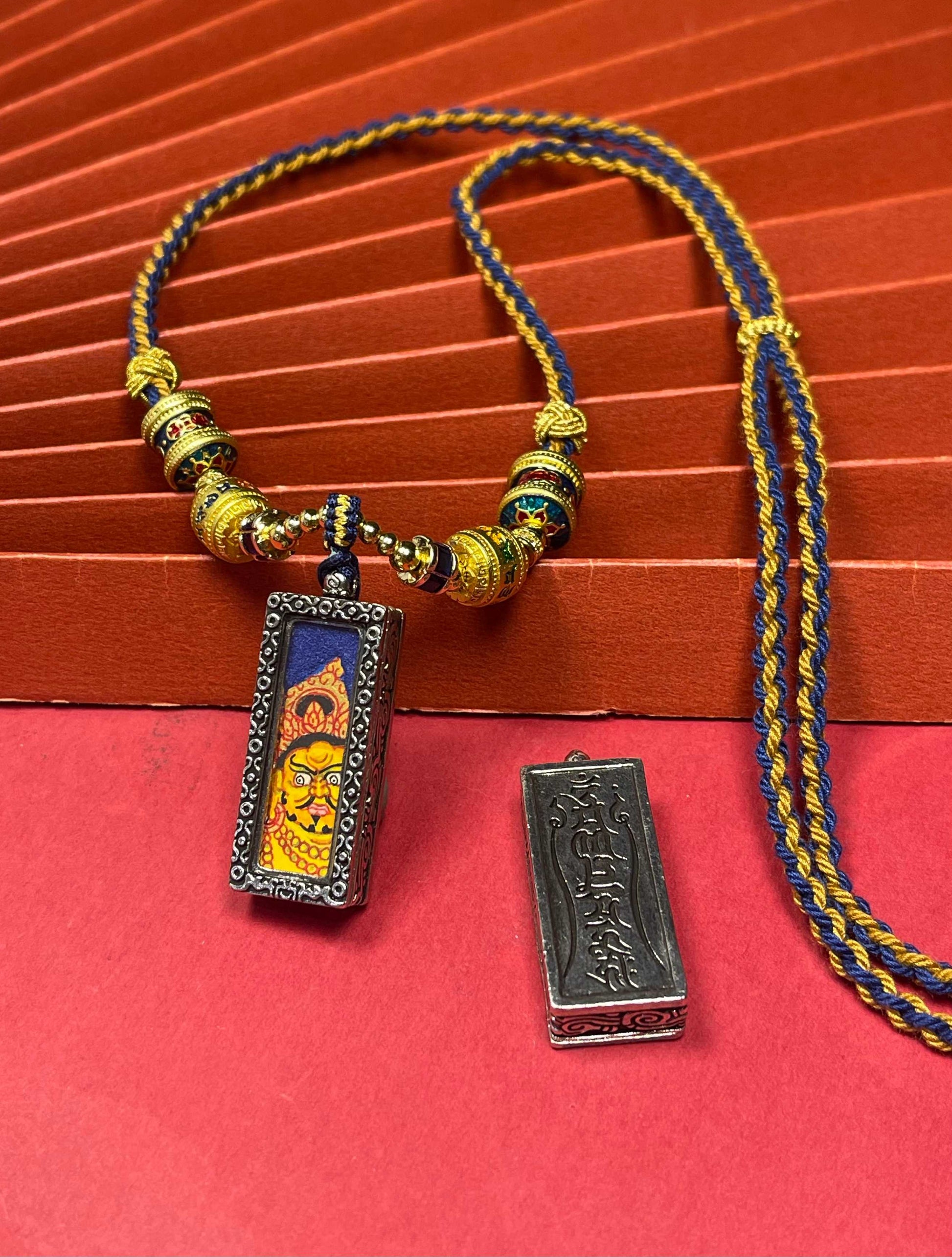  The Nepal Blue Base Bold Yellow Jambhala Thangka pendant with silver copper casing displayed as a sacred amulet for prosperity, embodying Buddhist tradition.
