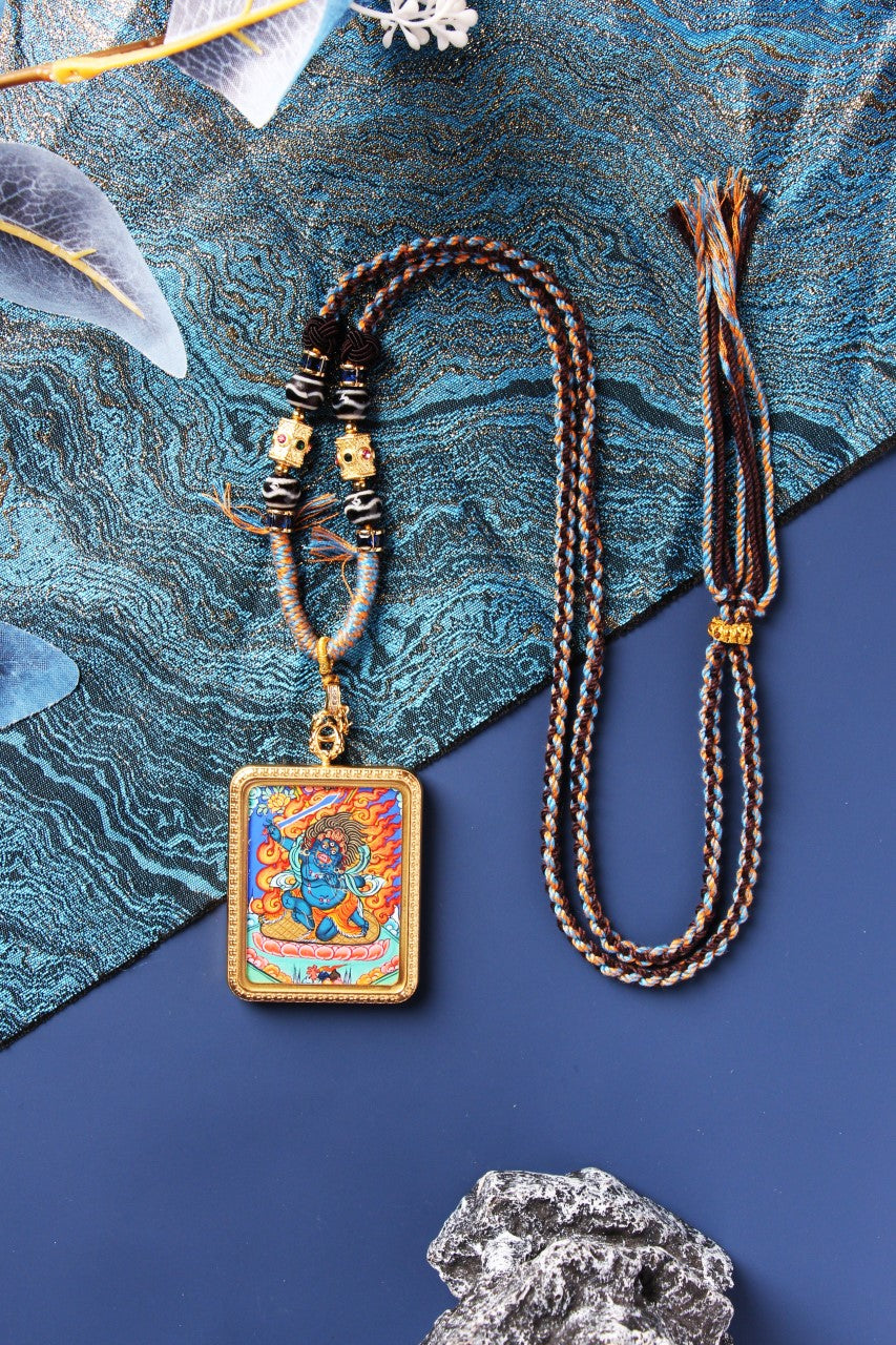A Rebgong Thangka pendant displayed with a traditional Tibetan hand-woven beaded cord and textured blue background.